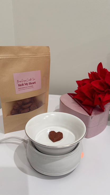 Photo of chocolate heart shaped wax melt in a wax melter. In the background are red flowers, a pink heart box, and the product packaging. 