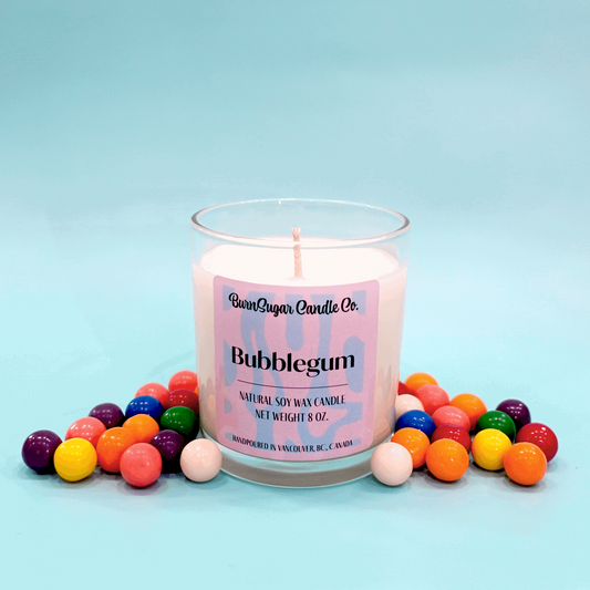 The image depicts a candle labeled "Bubblegum" from BurnSugar Candle Inc. It is an 8 oz natural soy wax candle, hand-poured in Vancouver, BC, Canada. The candle is surrounded by colorful gumballs, set against a light blue background. 