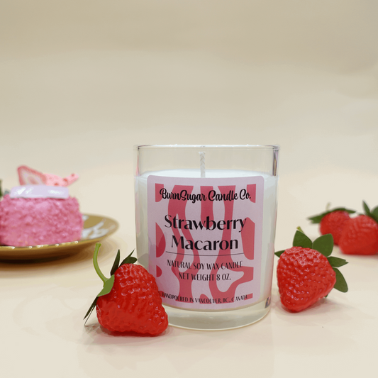 The image shows a candle labeled "Strawberry Macaron" from BurnSugar Candle Inc. It is an 8 oz natural soy wax candle, hand-poured in Vancouver, BC, Canada. The scene is styles with fresh strawberries in the foreground and a pink strawberry dessert on a gold plate in the background, set against a light beige backdrop. 